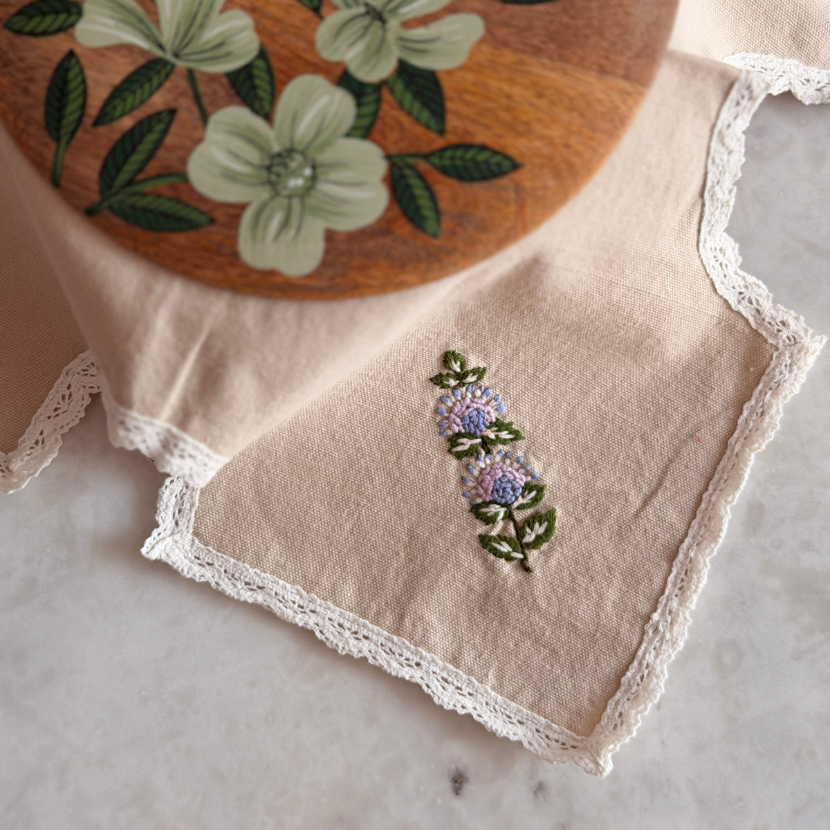 A close-up of a cream linen napkin with white lace trim and a small embroidered floral motif, partially folded over a round wooden board decorated with painted flowers.
