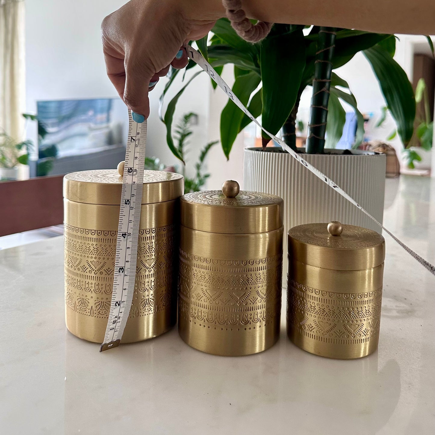 Three gold decorative canisters on a surface with a measuring tape, against a blurred indoor background.