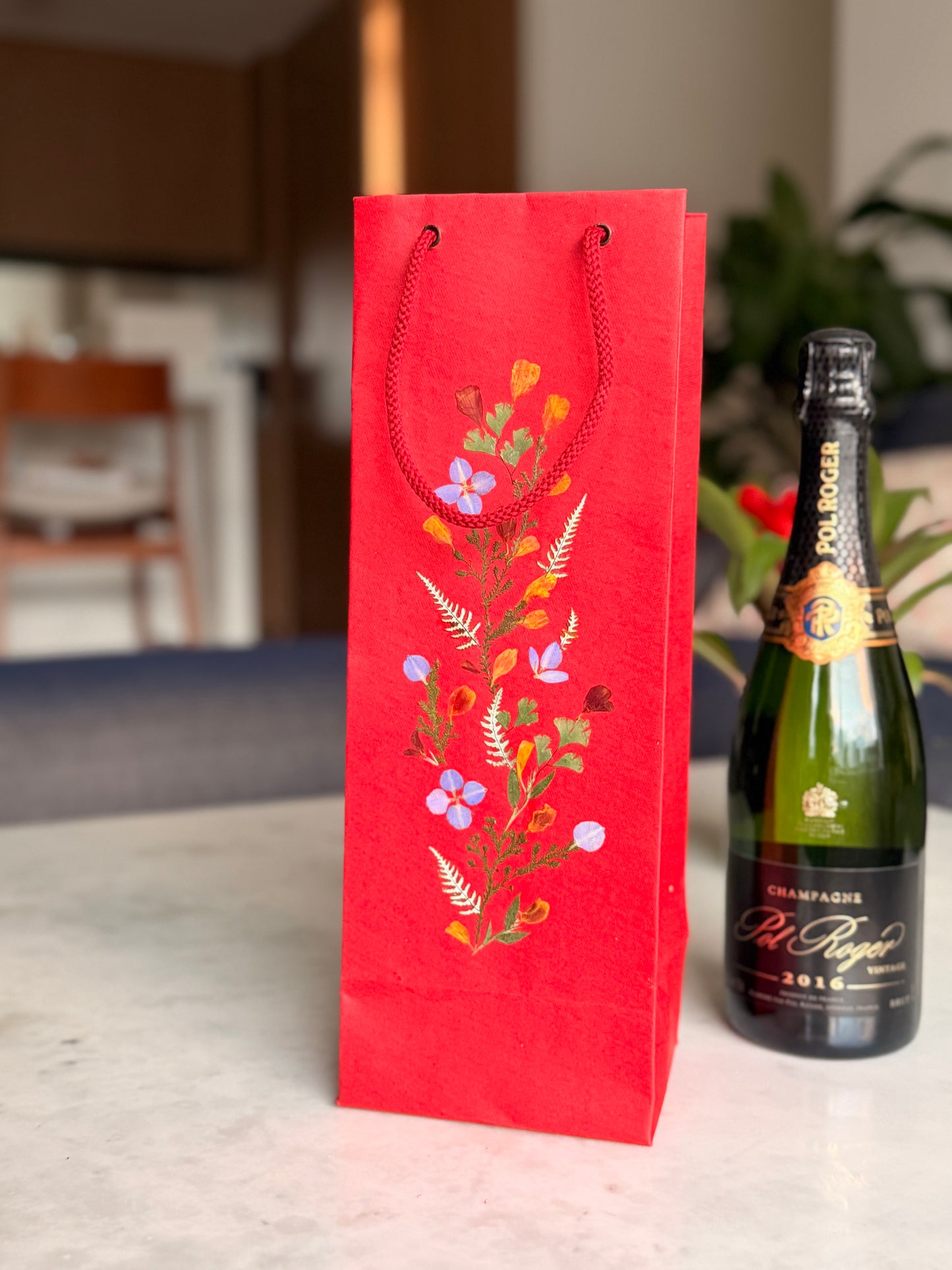 Red floral gift bag next to a bottle of champagne on a table.