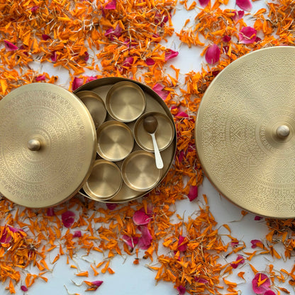 A brass spice container with a round lid, shown open with six small round spice jars inside and a small spoon, surrounded by scattered orange flower petals and pink rose petals on a light blue background.