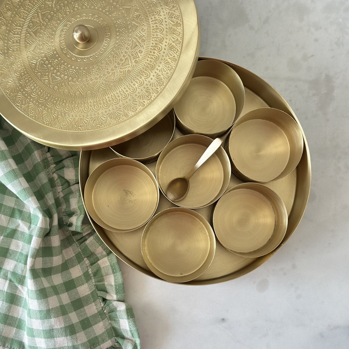 Brass masala dabba with lids on a marble surface next to a green checkered cloth