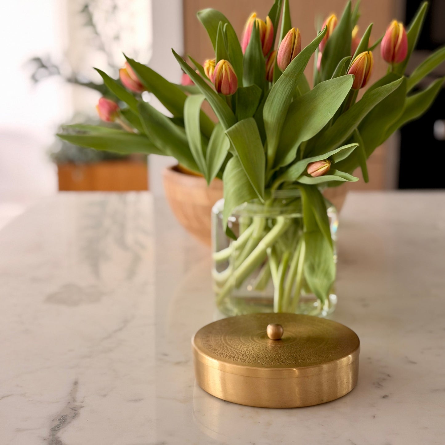 A round, brushed gold metal container lid with a small central knob sits on a white marble countertop in front of a glass vase holding tulips with pink and yellow petals.