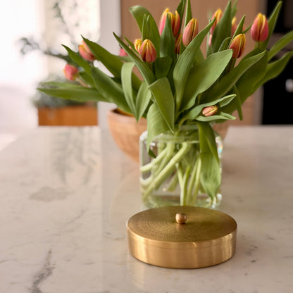 A round, brushed gold metal container lid with a small central knob sits on a white marble countertop in front of a glass vase holding tulips with pink and yellow petals.