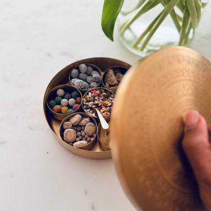 A brass spice container with multiple compartments filled with nuts, seeds, and colorful candies, shown on a white marble surface with a hand lifting the lid.