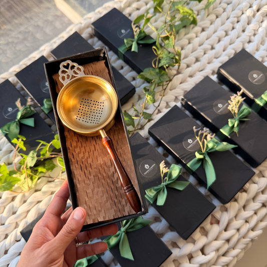 Brass Tea strainer in a box held by a hand with black boxes and greenery in the background