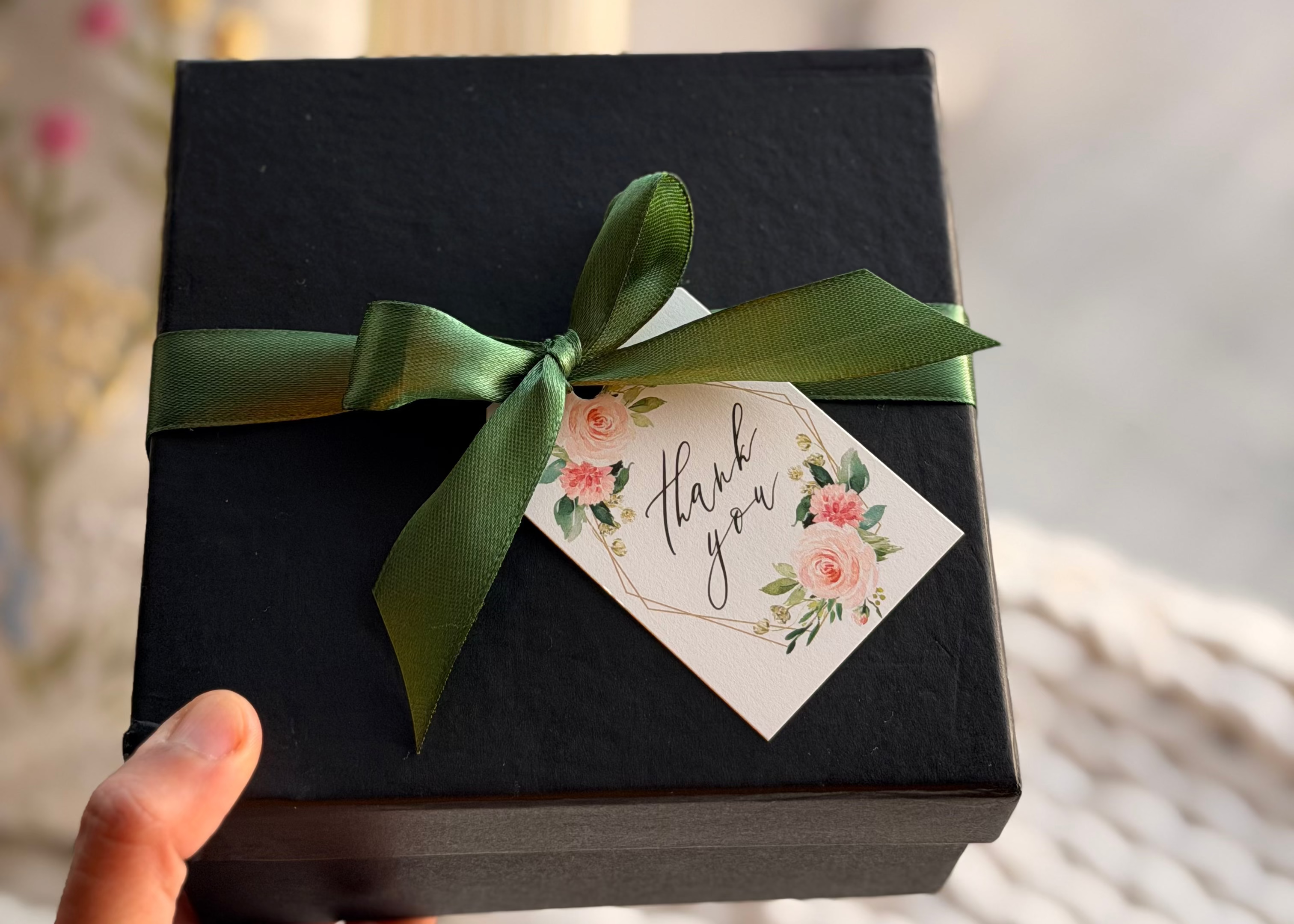 Black gift box with a green ribbon and floral tag held by a hand against a blurred background