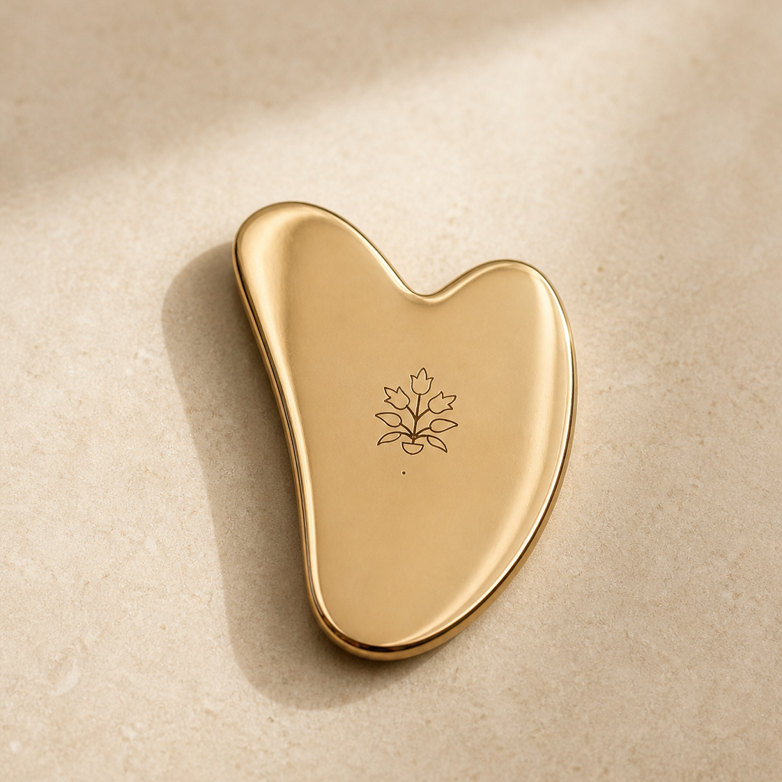Kansa gua sha by Vaarsa London, polished gold heart shape with engraved leaf mark, on cream stone