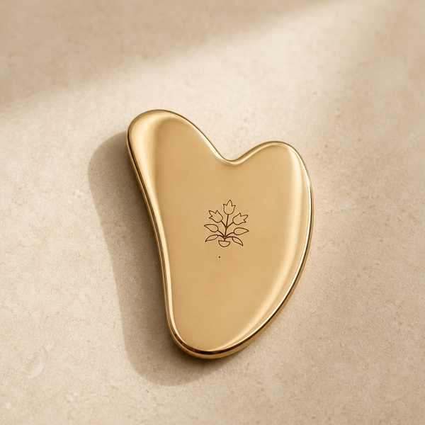 Kansa gua sha by Vaarsa London, polished gold heart shape with engraved leaf mark, on cream stone