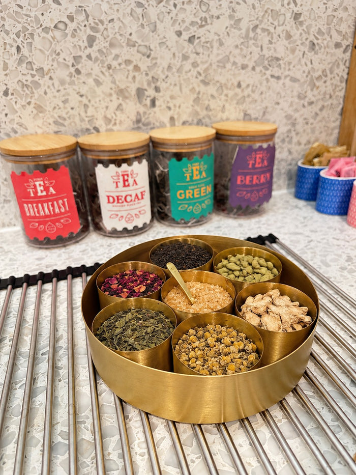 Brass spice box set up as a chai station with loose tea, sugar, cardamom, dried ginger, rose and chamomile on a kitchen counter