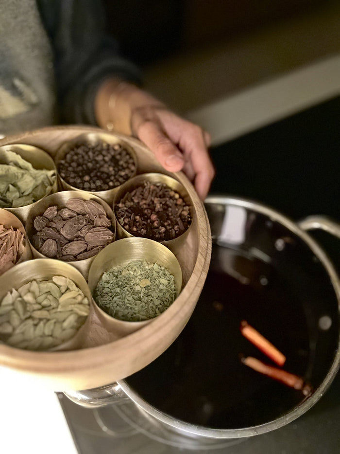 Brass spice box of whole spices held over a pot of mulled wine with cinnamon sticks