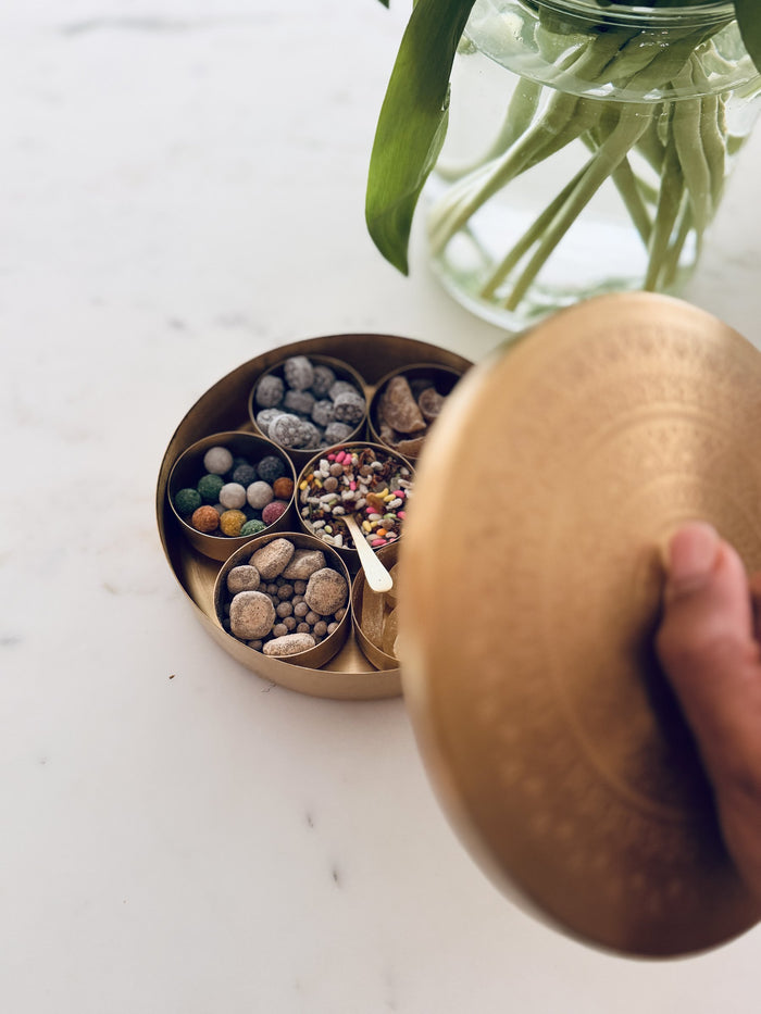Hand lifting the etched brass lid off a spice box filled with mukhwas and sweets, tulips in a glass vase behind