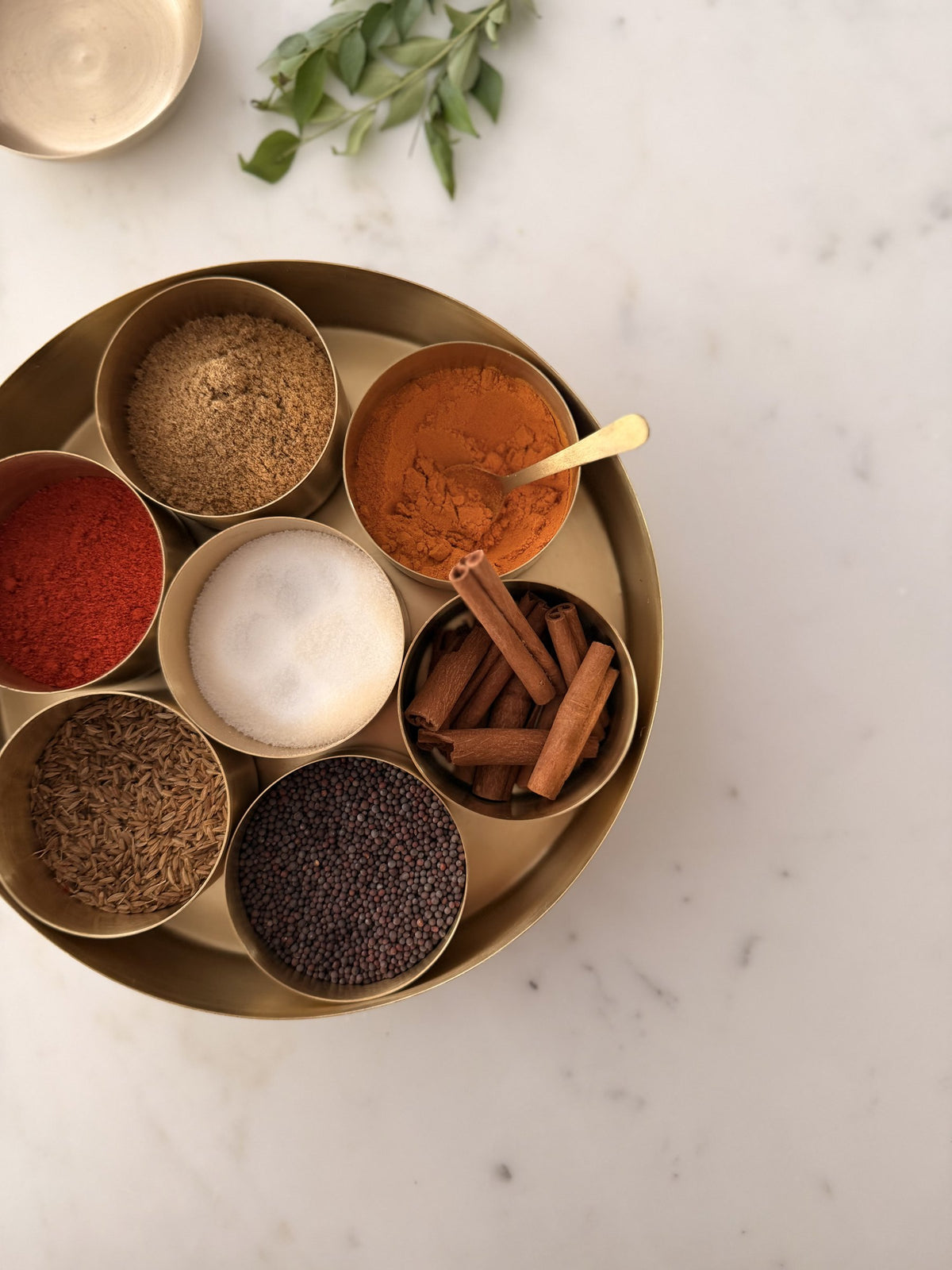 Brass masala dabba from above with seven bowls of chilli, turmeric, salt, cinnamon, cumin and mustard seed, a brass spoon and curry leaves on marble