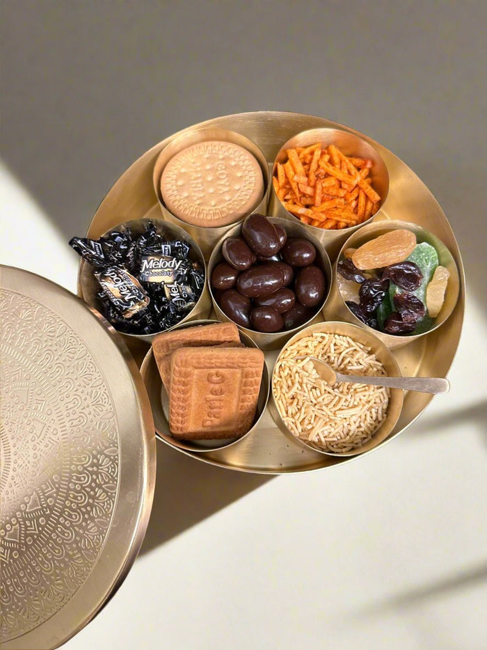 Brass spice box from above filled with Parle-G and Marie biscuits, namkeen, chocolate almonds, toffees and dried fruit, the etched lid beside it